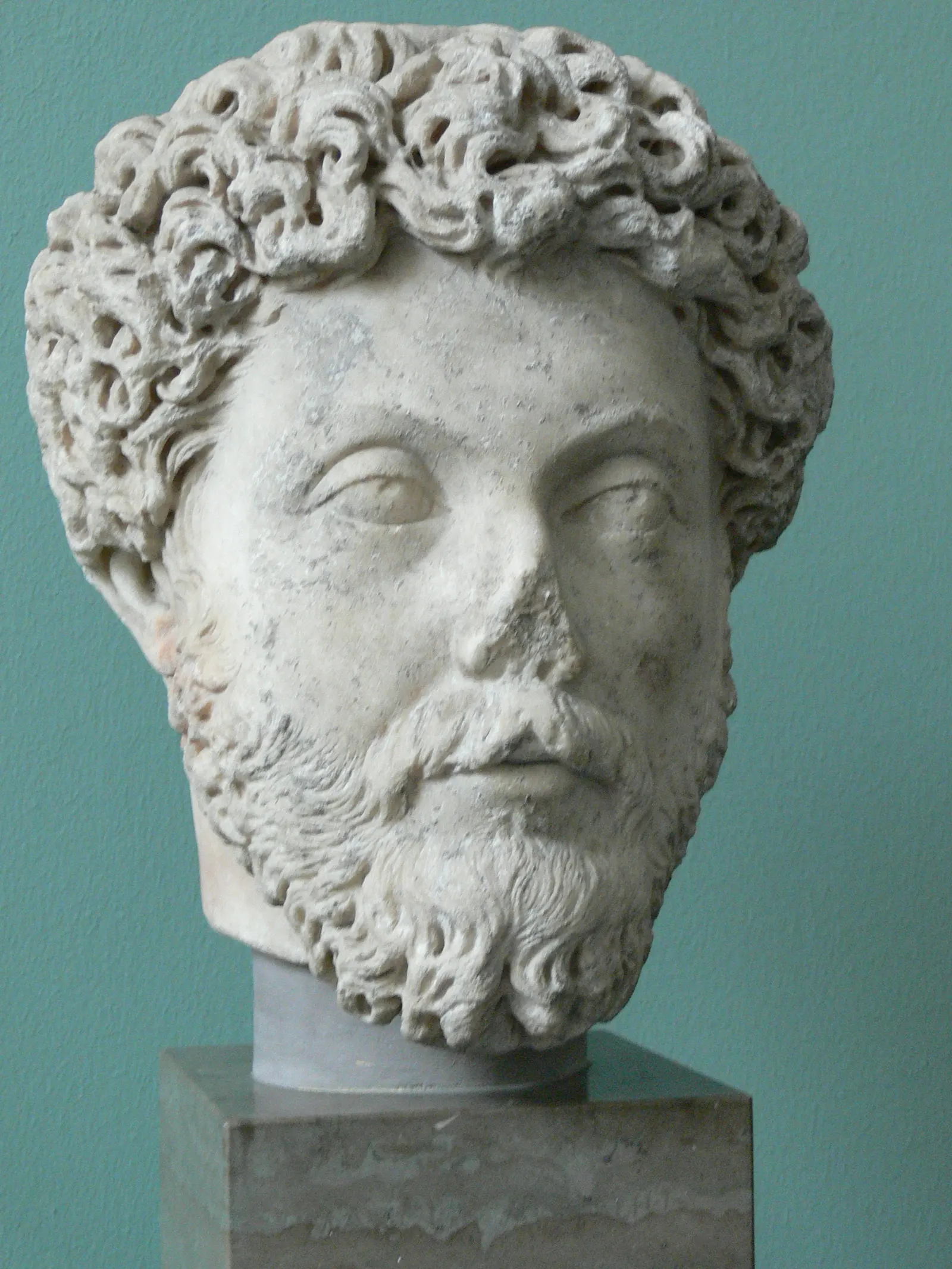 Portrait bust of Marcus Aurelius, curly hair and beard, on a stone pedestal against a green museum wall