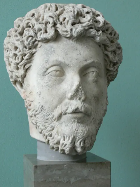 Portrait bust of Marcus Aurelius, curly hair and beard, on a stone pedestal against a green museum wall